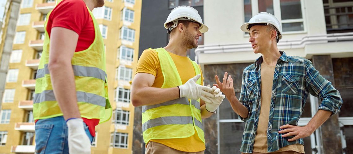 Communication. Engineer in safety helmet and plaid shirt smiling gesture and rude and two workers in gloves and vests listening at construction site during day